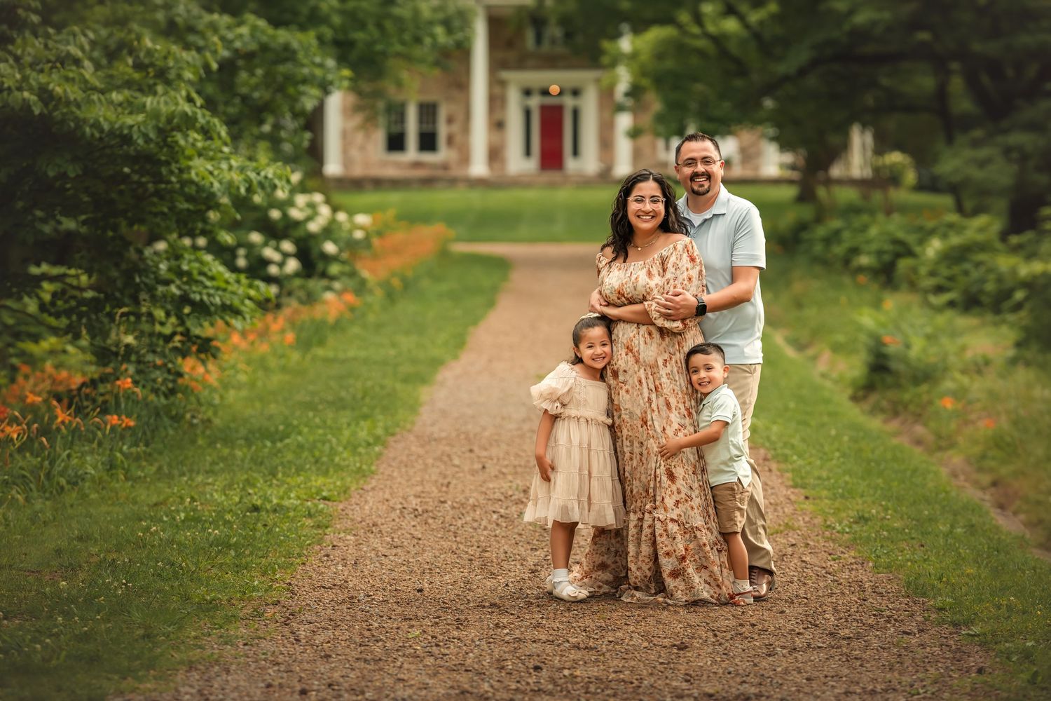 Family of four poses on a gravel path before a white house surrounded by lush greenery.