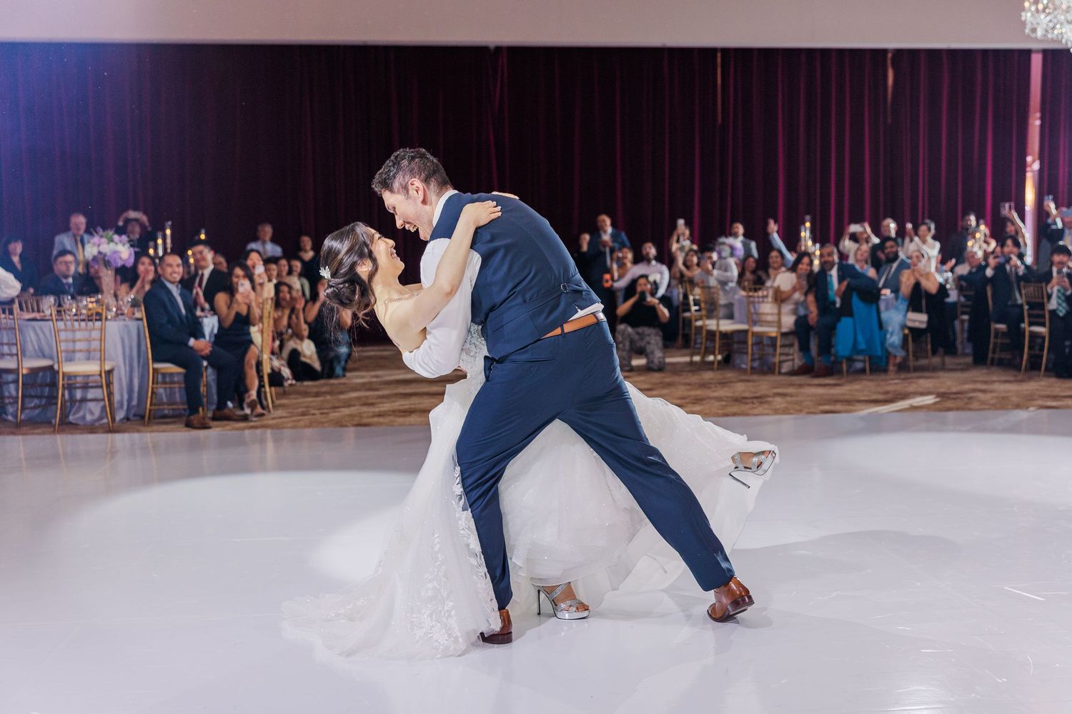 Bride and groom share romantic first dance dip on white dance floor at elegant wedding reception.