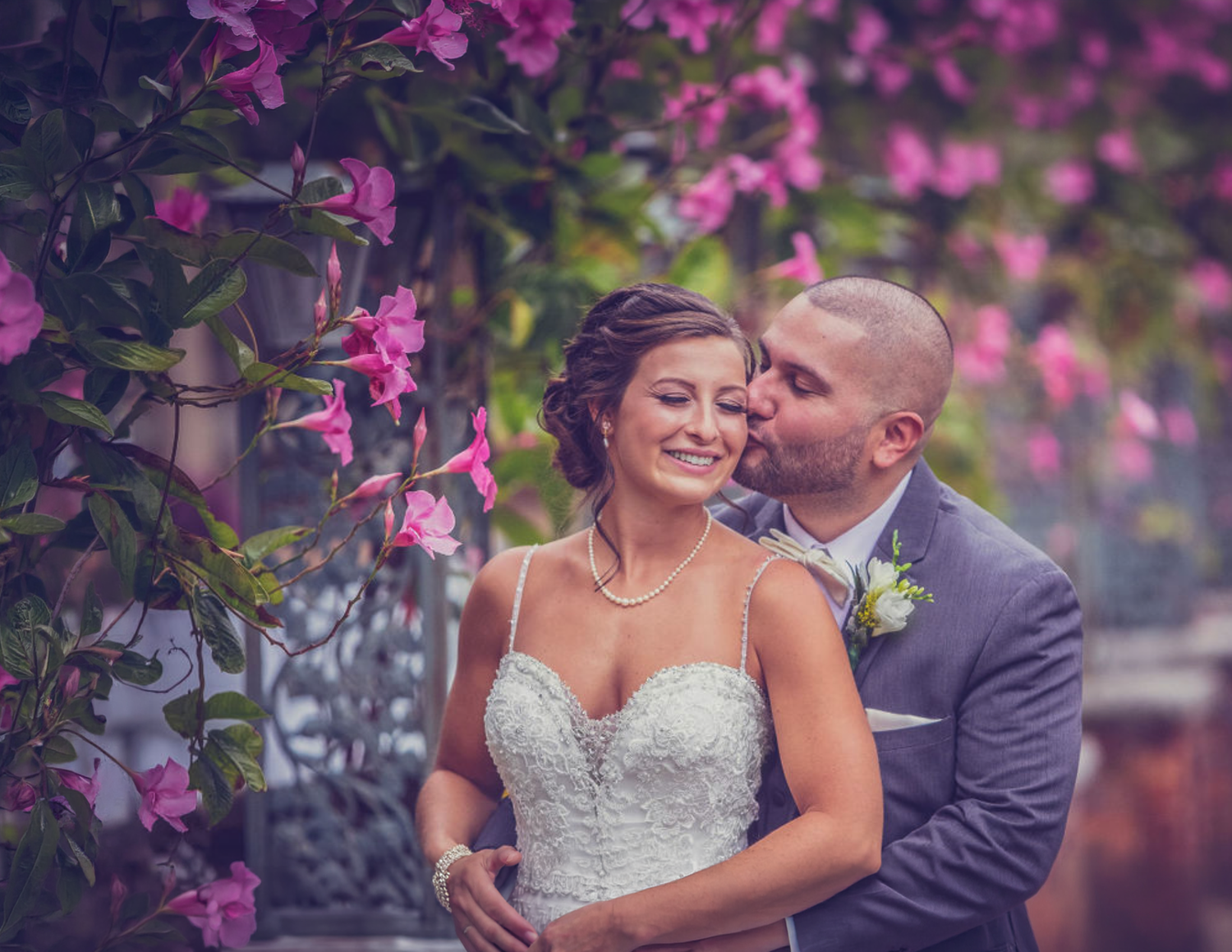 Bride and groom embrace under blooming pink flowers in a romantic outdoor wedding portrait.