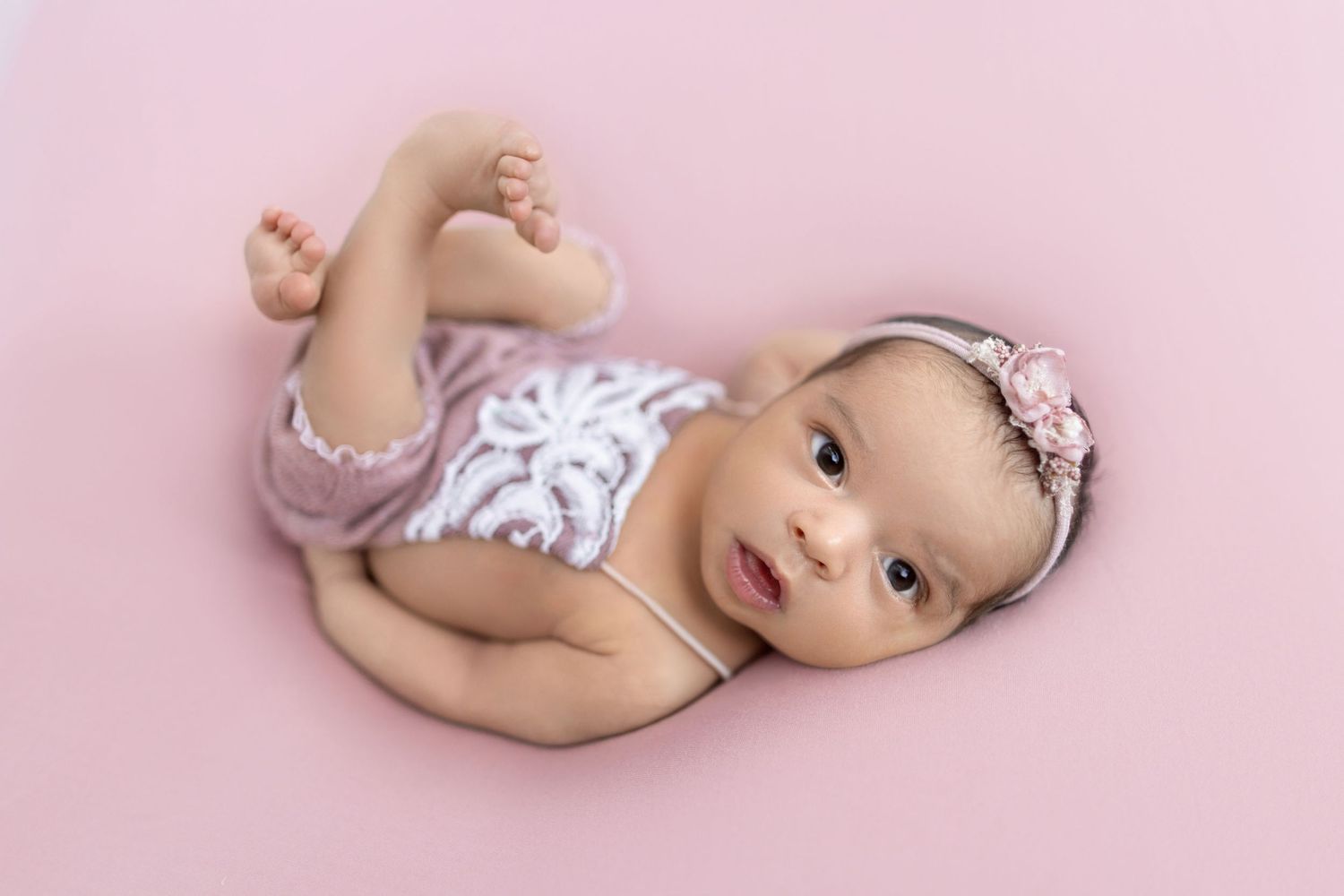 Newborn baby girl in pink lace romper and floral headband lying on pink backdrop.