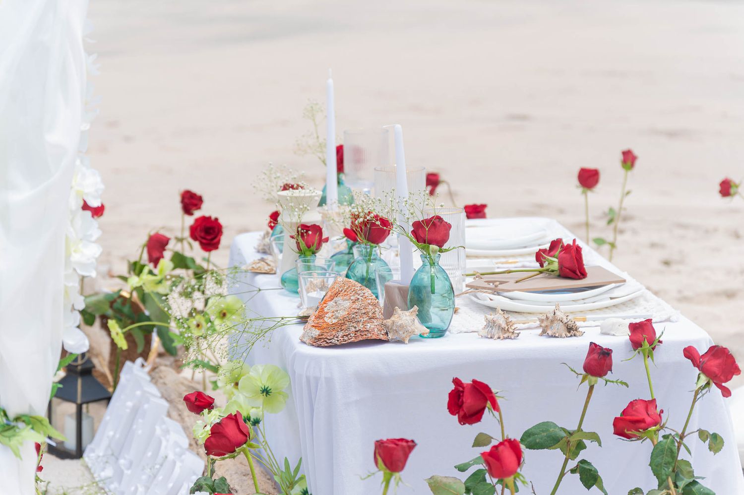 Romantic beach table setting with red roses, white candles, and turquoise vases on a white tablecloth.