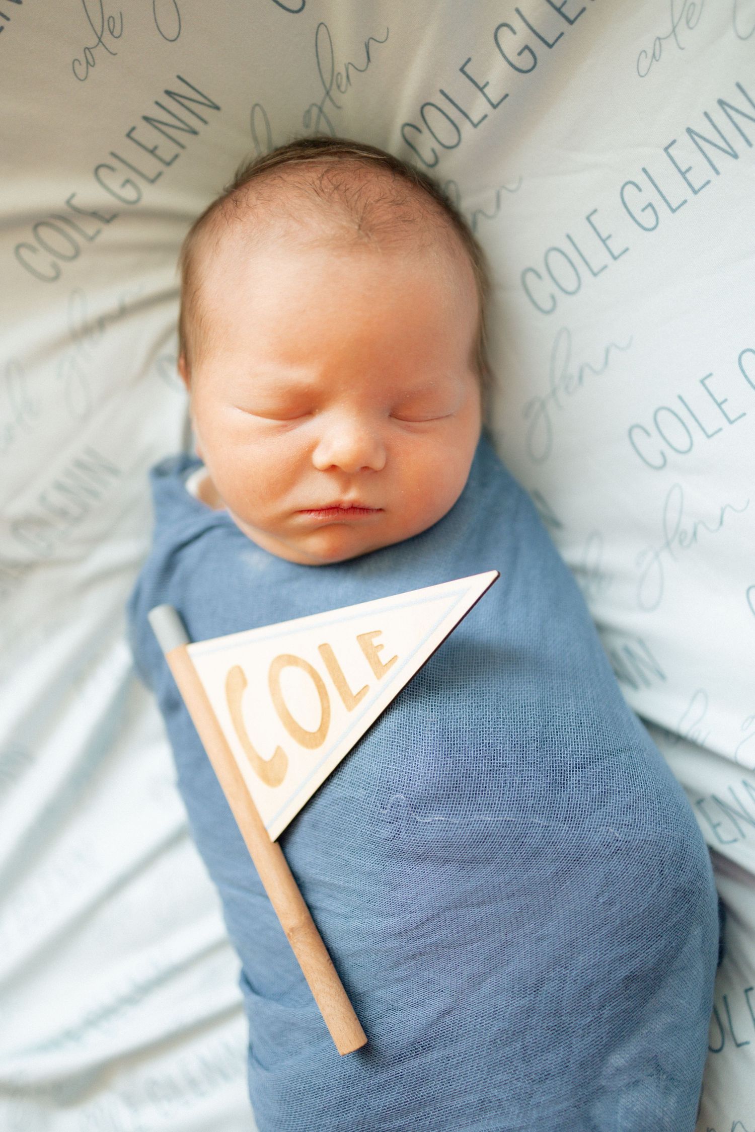 Newborn baby named Cole swaddled in blue wrap, sleeping peacefully on a personalized blanket with a small pennant flag.