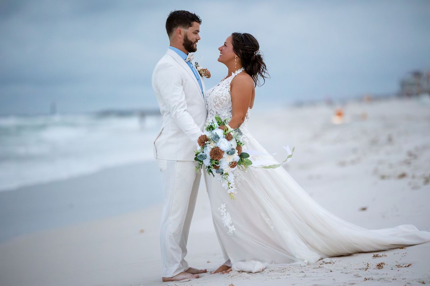 Couple sharing a romantic stare on a beach, man in white suit and woman in white dress at dusk.