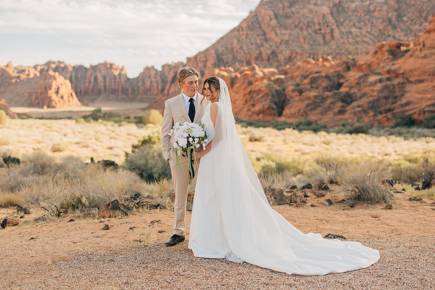 Bride and groom pose in desert landscape with red rock formations at golden hour wedding photo.