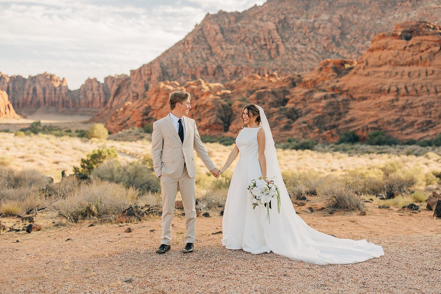 Bride and groom holding hands in desert landscape with red rock formations at sunset.
