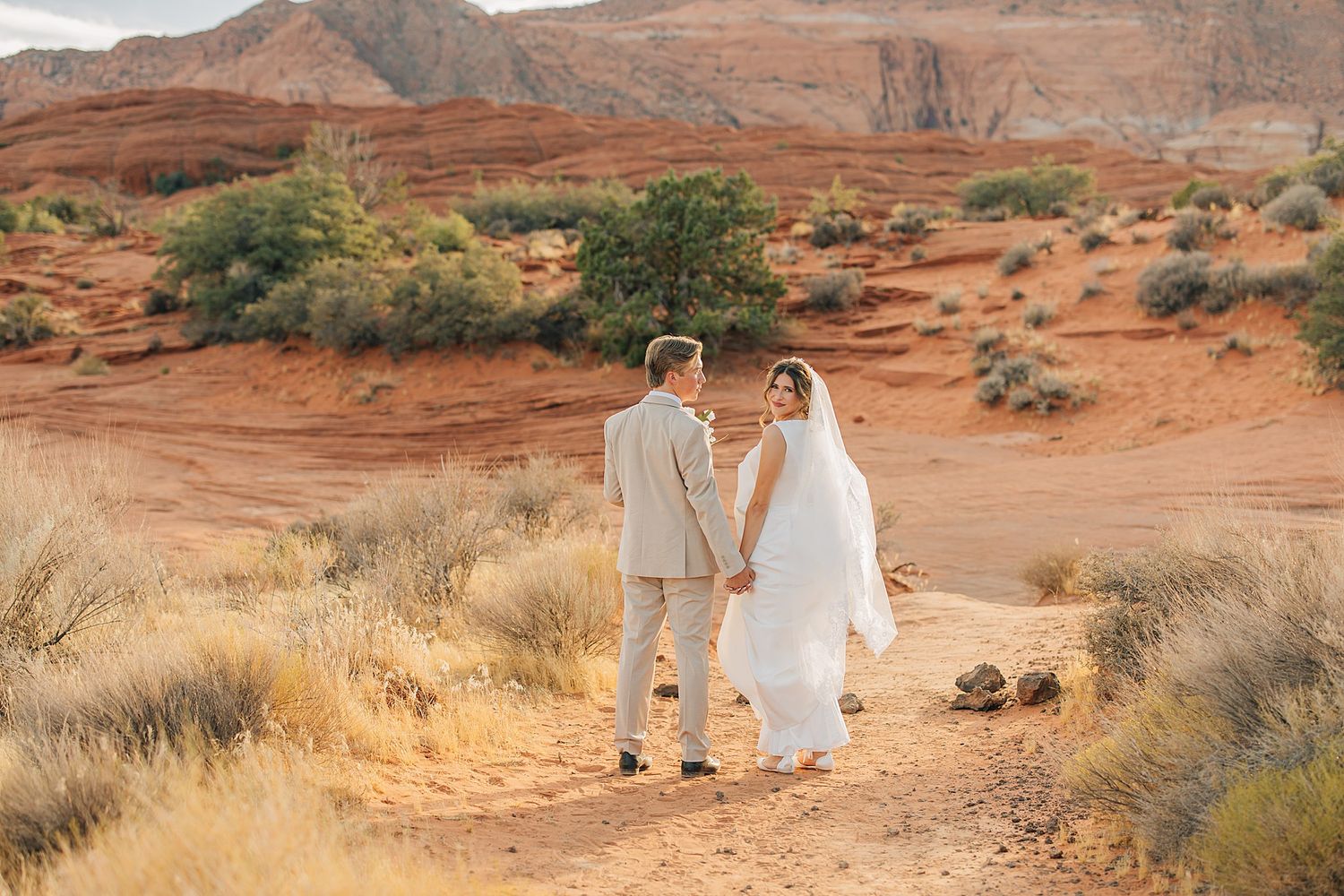 Bride and groom hold hands in stunning red rock desert landscape, bride in white gown with veil, groom in tan suit.