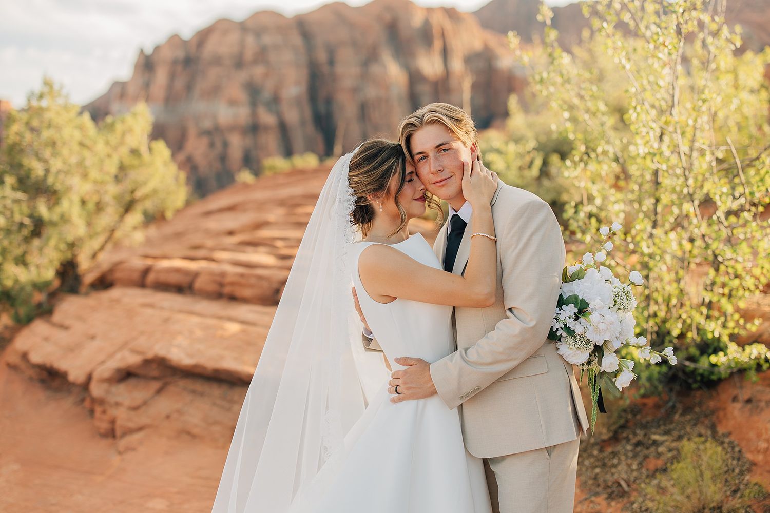 Bride and groom embrace at Sedona red rock backdrop, she in white gown with veil, he in tan suit holding bouquet.