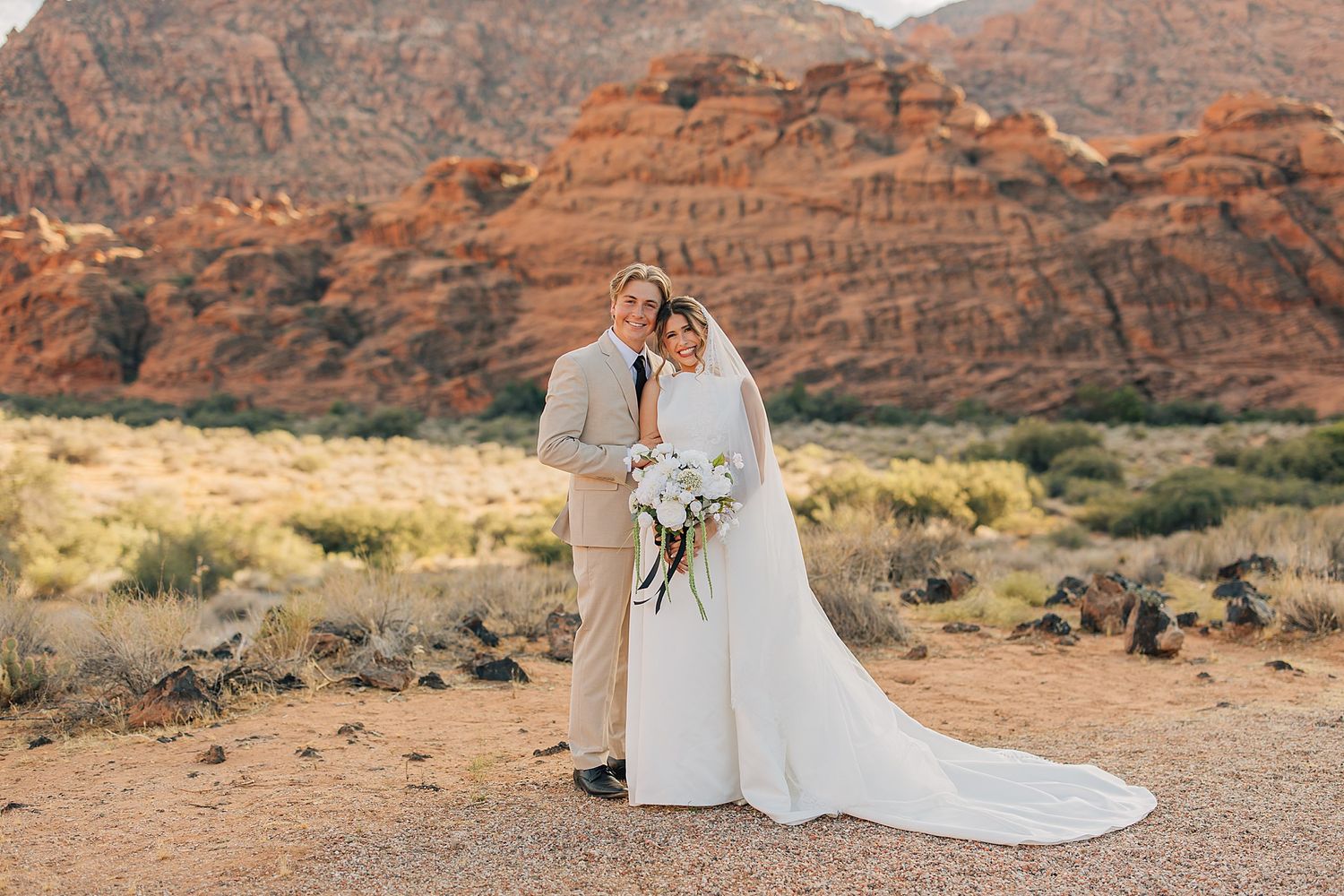 Bride and groom pose together at Red Rock Canyon desert wedding with white gown, bouquet, and tan suit.