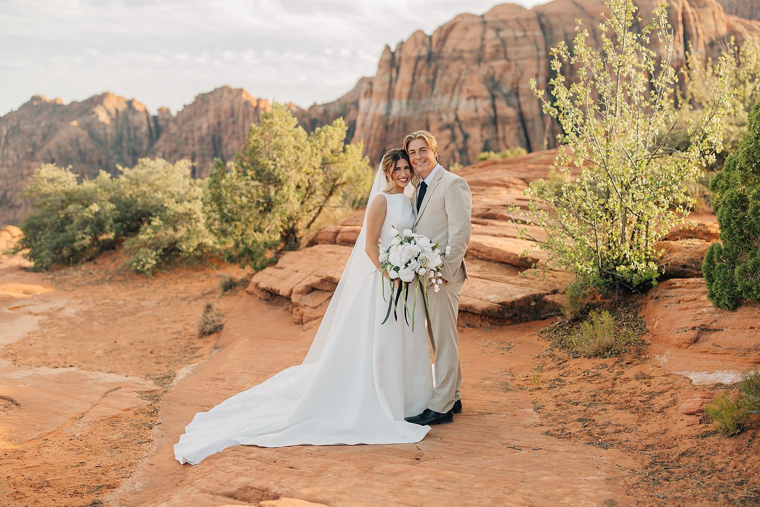 Bride and groom pose together in white wedding attire against red rock canyon landscape at golden hour.