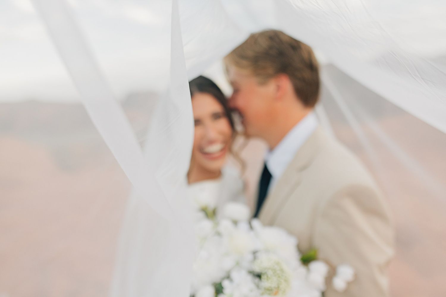 Blurred romantic wedding photo of bride and groom under white veil, bride holding white floral bouquet.
