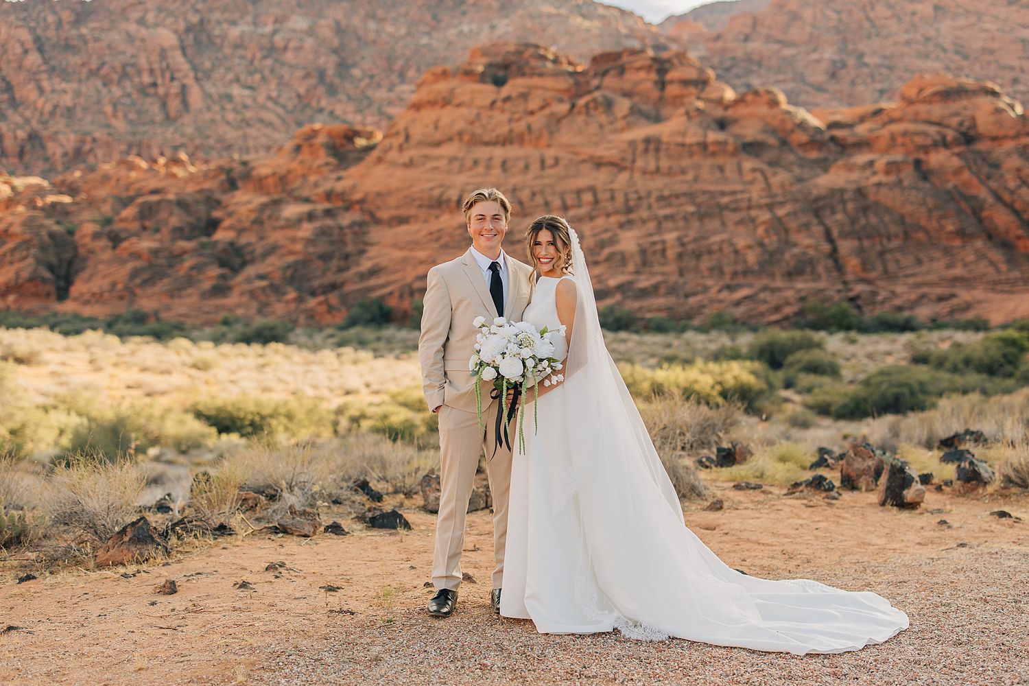 Couple poses in wedding attire before red rock canyon desert landscape, bride holding white bouquet.