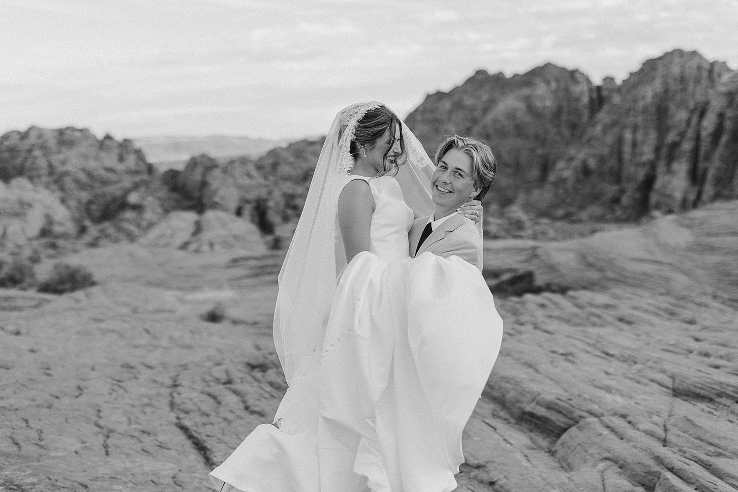 Black and white photo of groom carrying bride among dramatic rocky desert landscape.