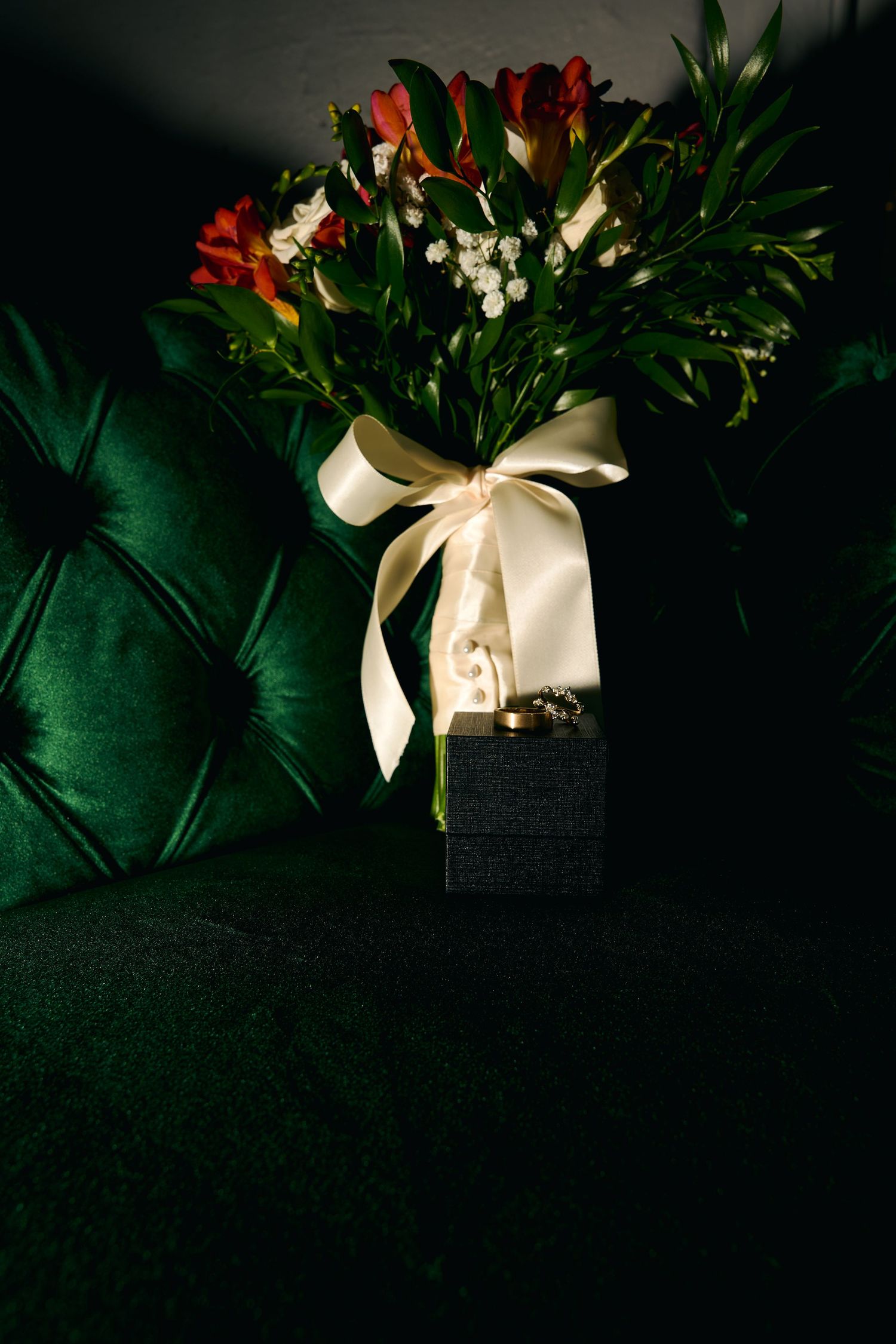 Elegant bridal bouquet of red alstroemeria with white ribbon bow resting on a dark gift box on green velvet sofa.
