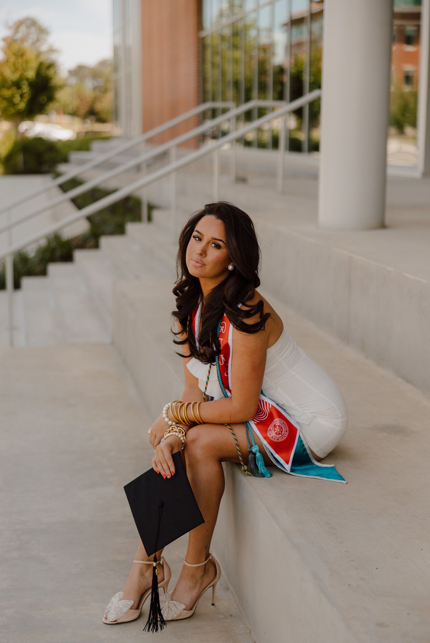 Young woman in white dress holding graduation cap sits on concrete steps, wearing gold bracelets and heels.
