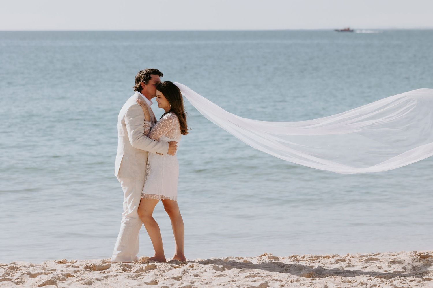 Couple kissing on a white sand beach in matching white outfits, with a flowing veil blowing in the ocean breeze.