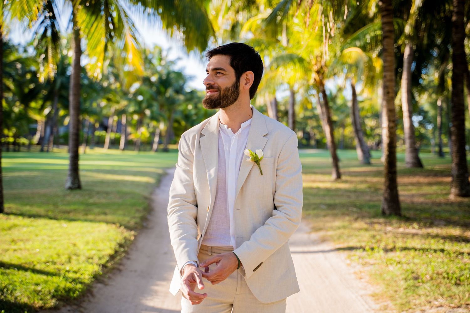 Groom in beige linen suit standing on tropical path surrounded by palm trees on sunny wedding day.