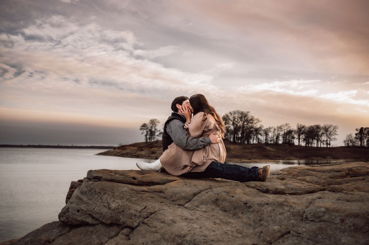 Couple sharing romantic kiss on rocky shoreline at sunset.