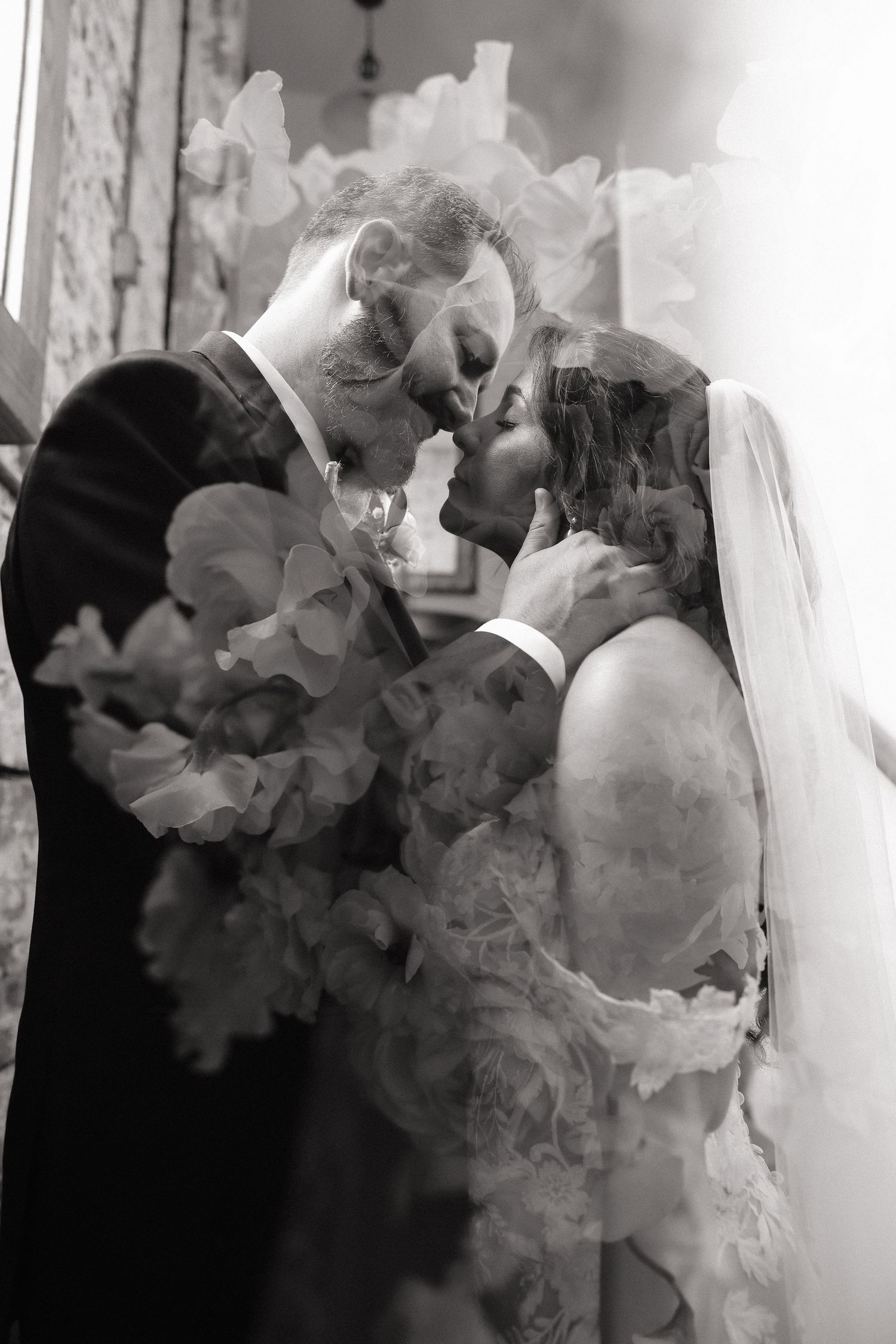 Black and white double exposure wedding photo of bride and groom nearly kissing, overlaid with soft floral blooms.