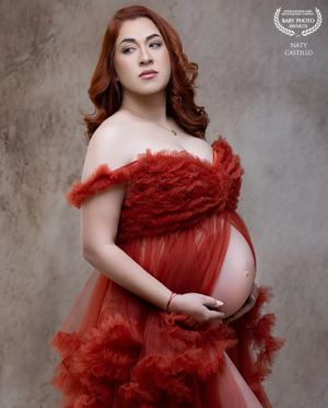 Elegant pregnant woman in red off-shoulder tulle gown cradling baby bump, professional maternity portrait.