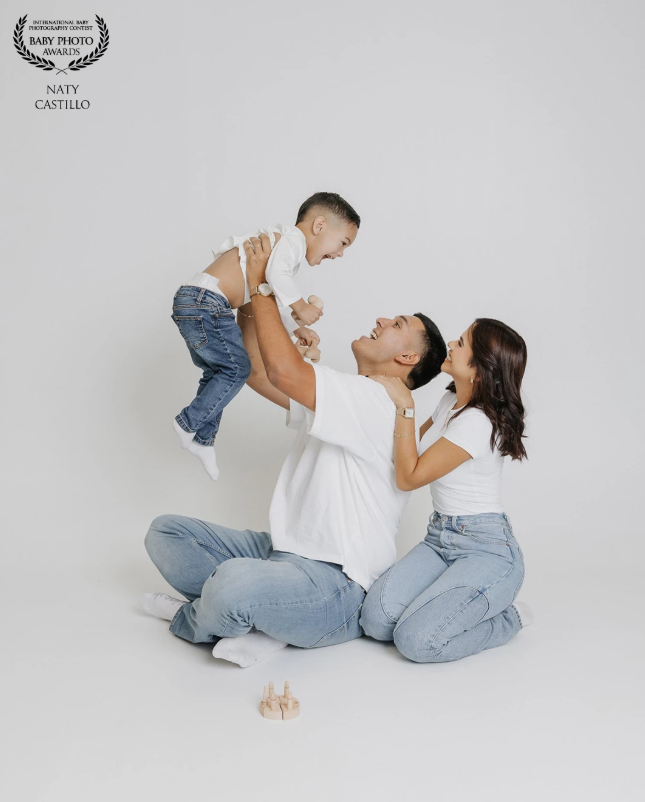 Happy family in white tops and jeans, parents lifting smiling baby in bright studio photo session.