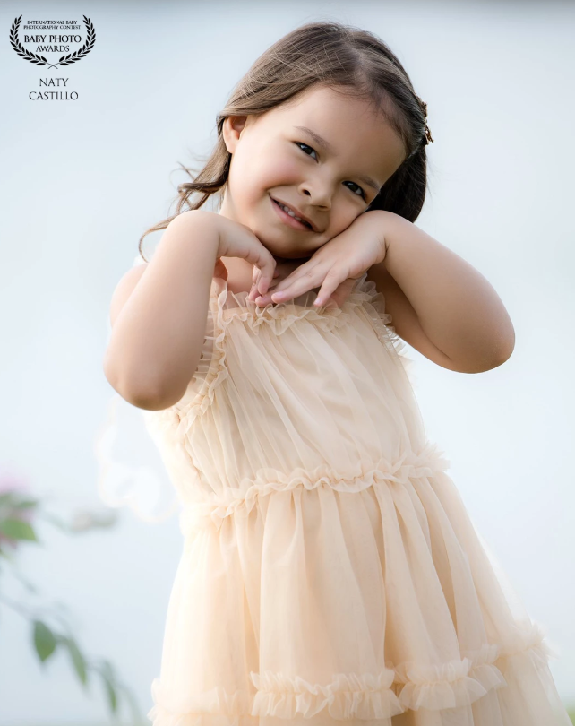 Smiling toddler girl in cream tulle dress posing with hands under chin, award-winning baby portrait photography.