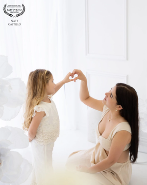 A mother and young daughter make a heart shape with their hands together in a bright white studio setting.