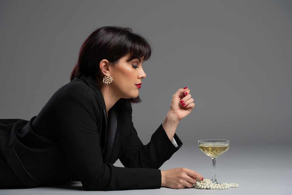 Elegant woman in black jacket lying down with pearl necklace and champagne glass on gray background.