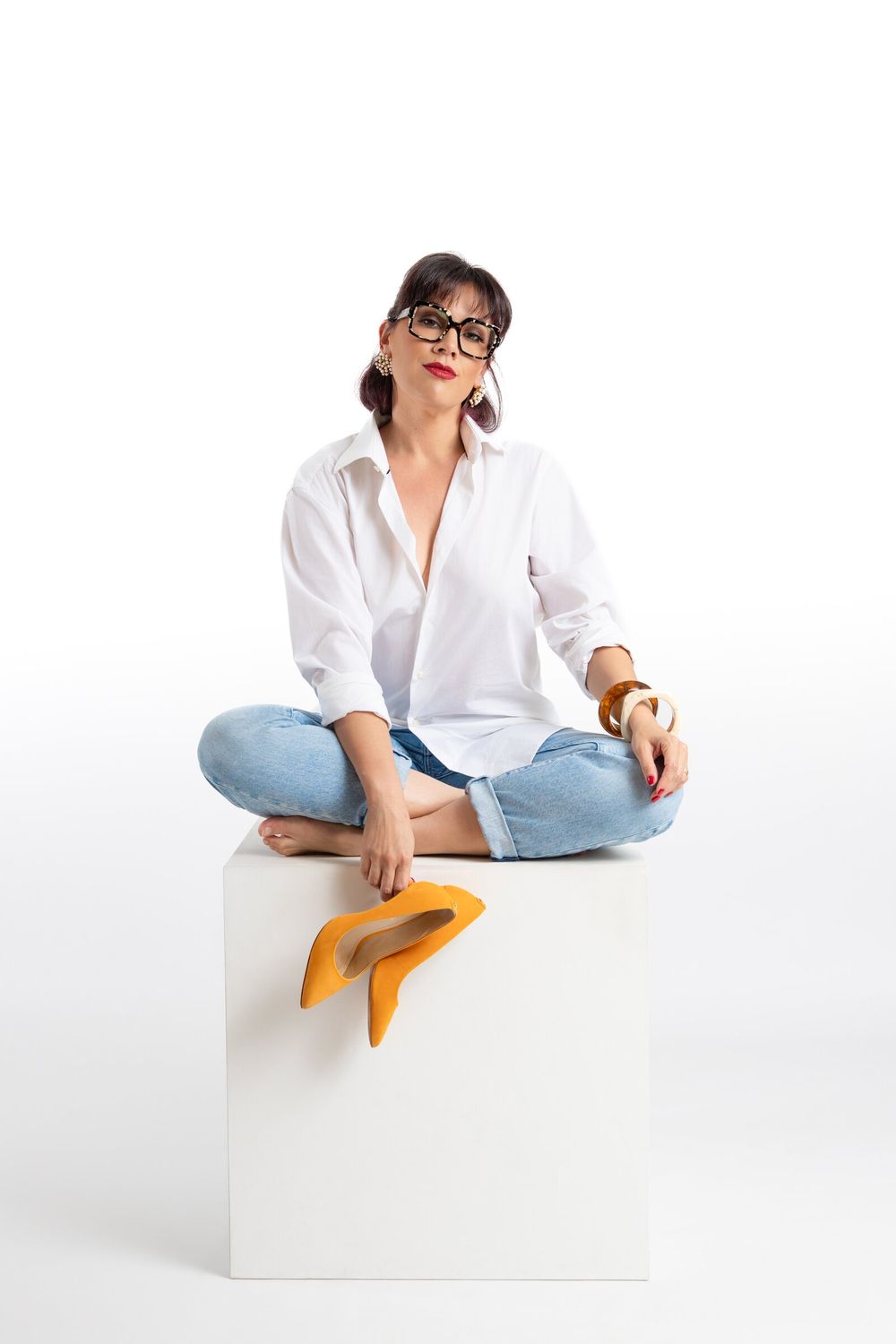 Woman in white shirt and jeans sitting cross-legged on white cube, holding yellow heels, wearing glasses.