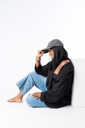 Woman in black top, jeans, and gray cap sits against white background, smiling downward with bracelets on wrist.