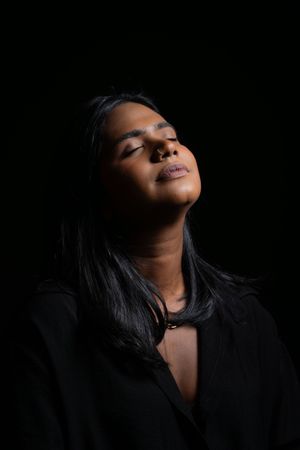 A South Asian woman in a black top tilts her head back with closed eyes against a dark background.