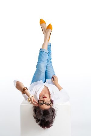 Woman in jeans and yellow heels lying on white cube with legs raised, wearing glasses and white shirt.