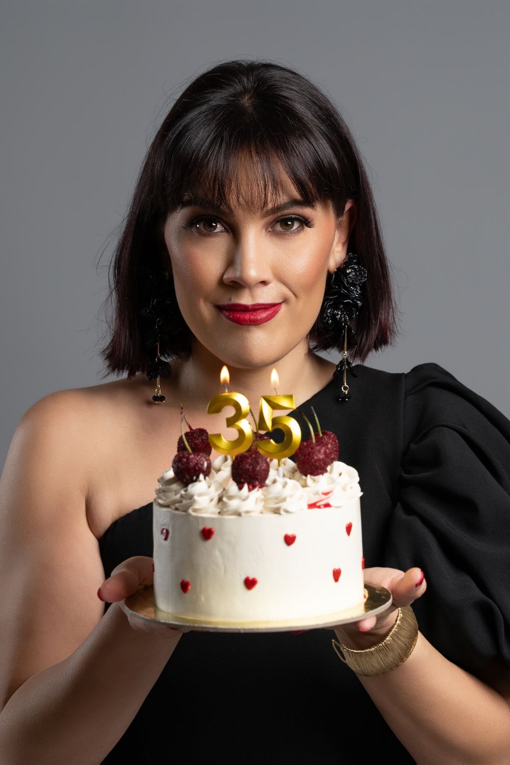 Woman in black dress holding a white birthday cake with gold number 35 candles and cherries on gray background.