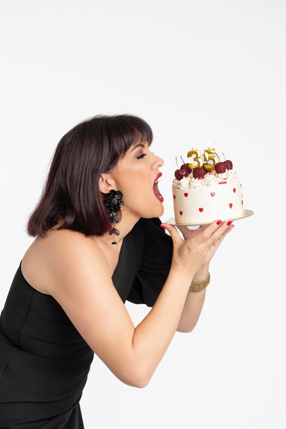 Woman in black dress playfully biting into a white birthday cake with number 35 candles and cherries on top.