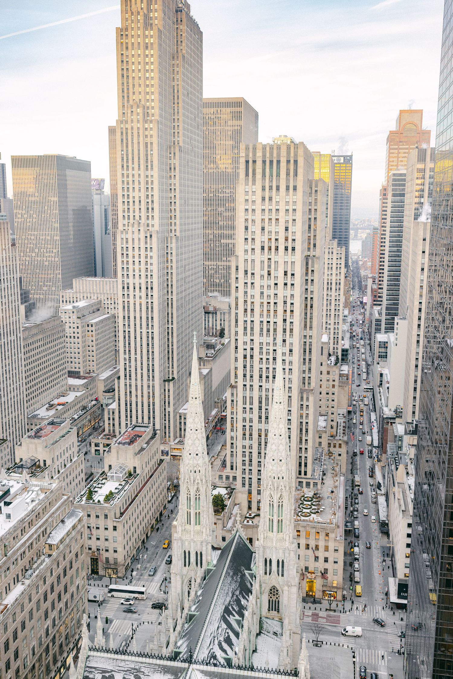 Aerial view of St. Patrick's Cathedral spires rising among Manhattan skyscrapers along a busy New York City avenue.