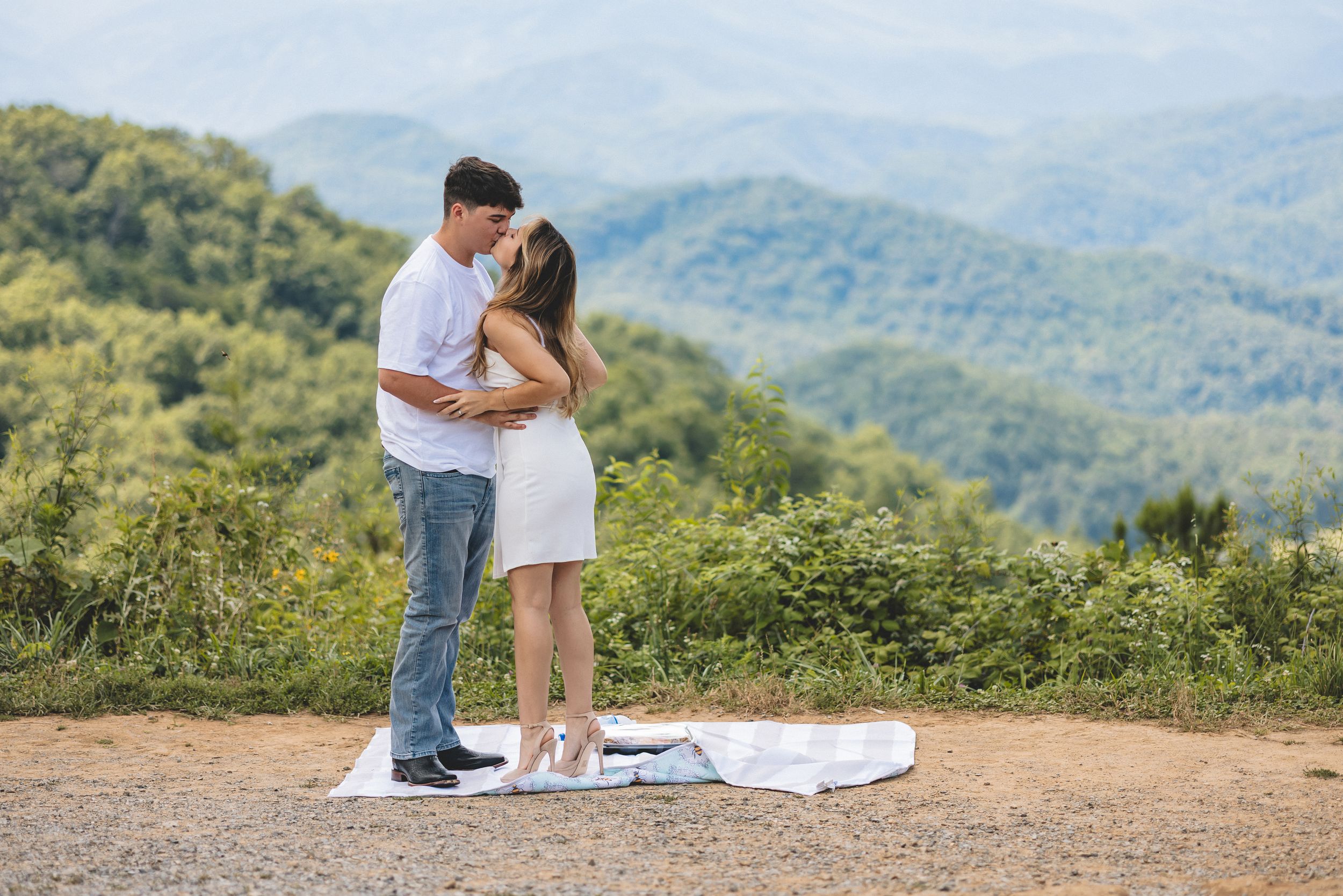 A couple shares a romantic moment on a mountaintop overlook with rolling hills and forests in the background.