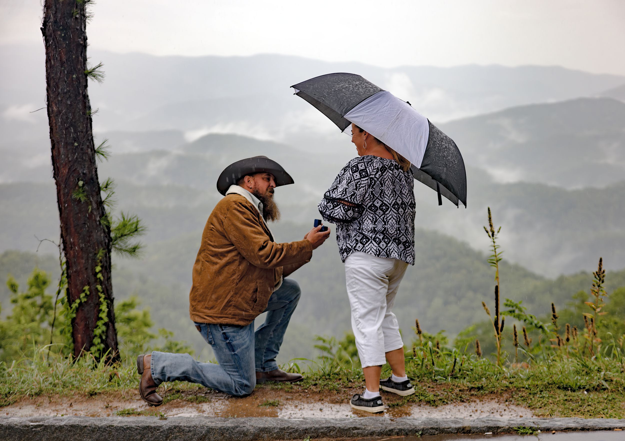Marriage proposal scene on rainy mountain overlook with umbrellas.