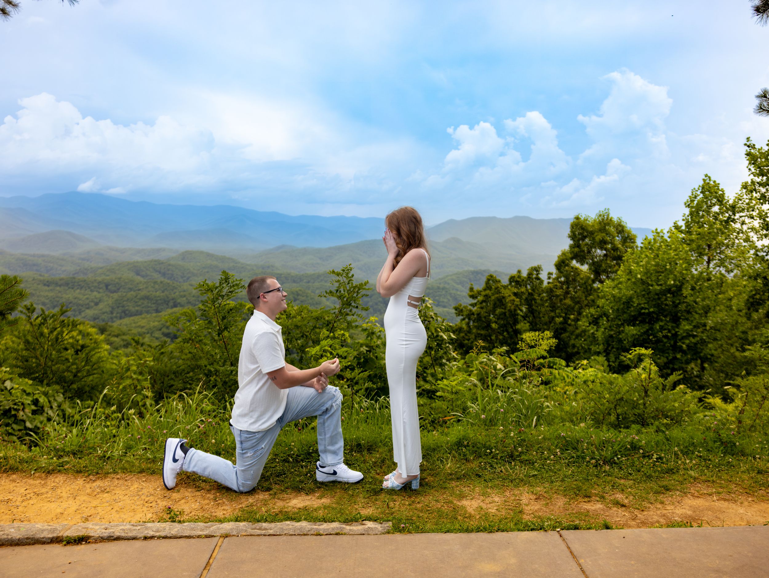 Man proposing on one knee to woman in white outfit at scenic mountain overlook with lush green hills and blue sky.