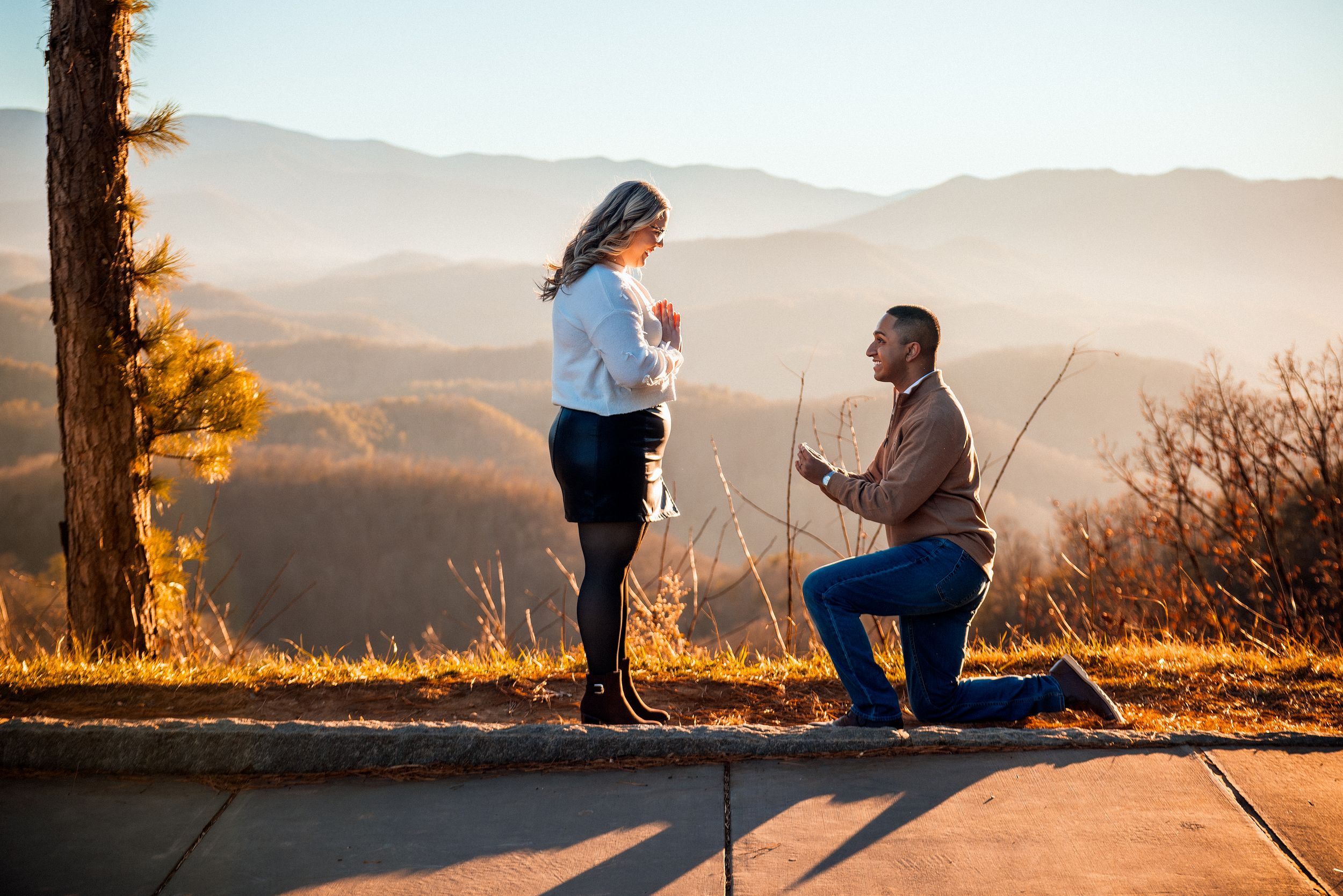 Two silhouettes stand on a mountain overlook during sunset as one person proposes to another.