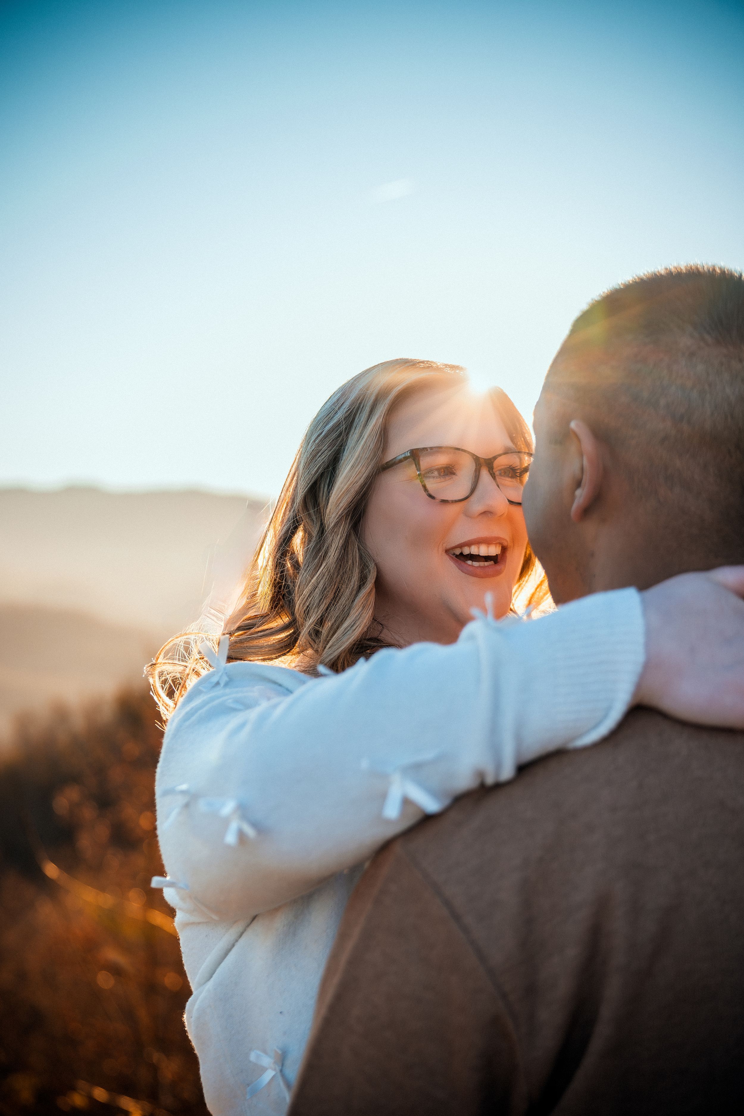 A joyful couple embraces outdoors during golden hour with sunlit mountains in the background.