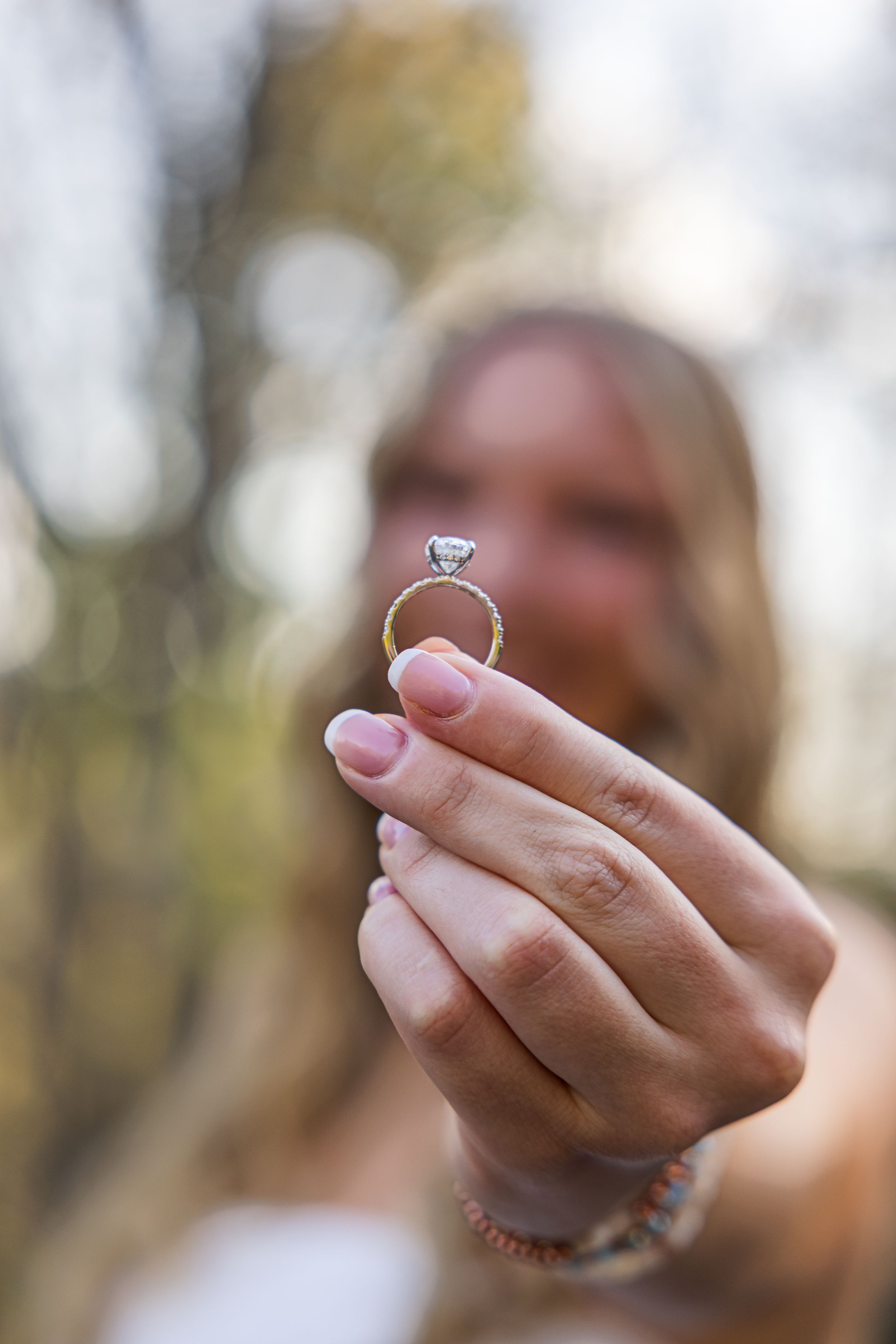 Woman holding up a diamond engagement ring outdoors with blurred bokeh background in natural light.
