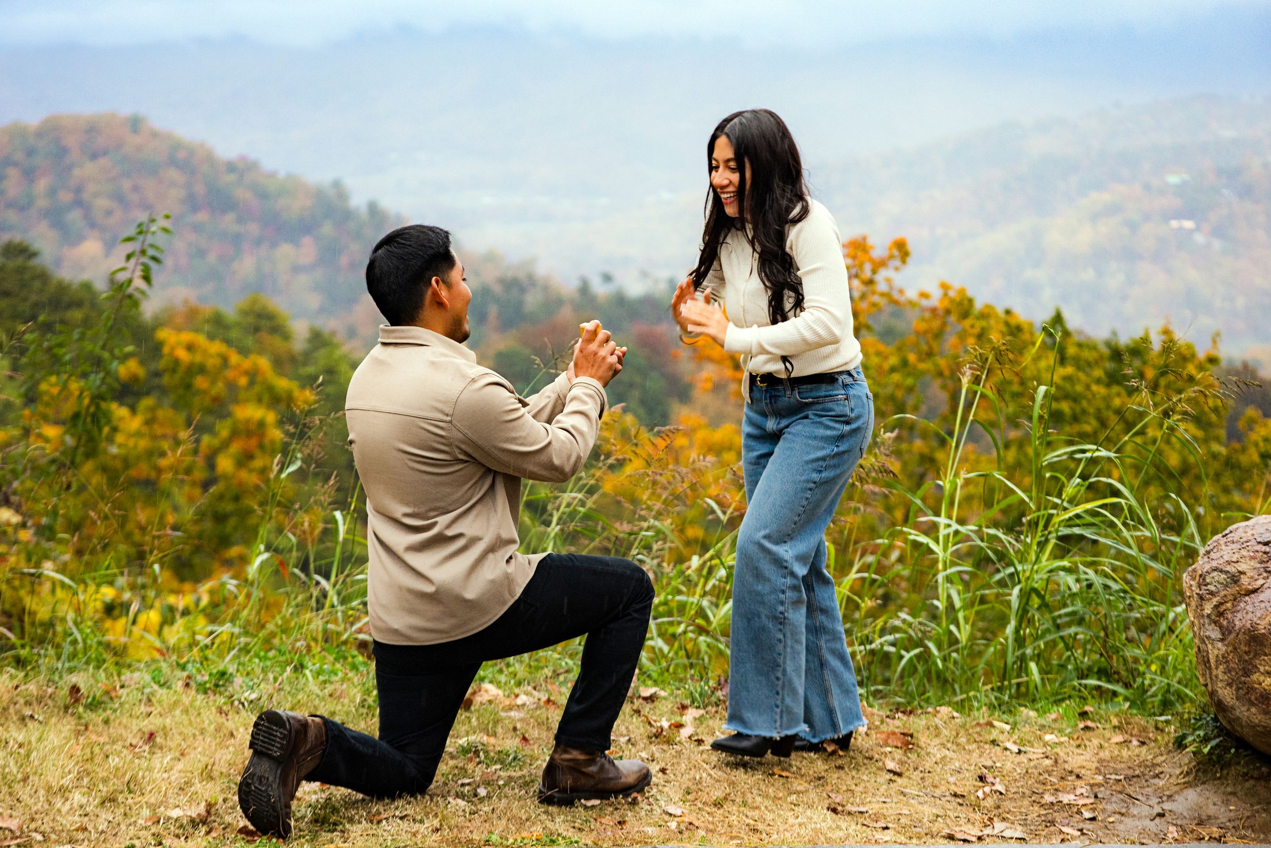 A couple shares a romantic proposal moment on a scenic mountain overlook during autumn.