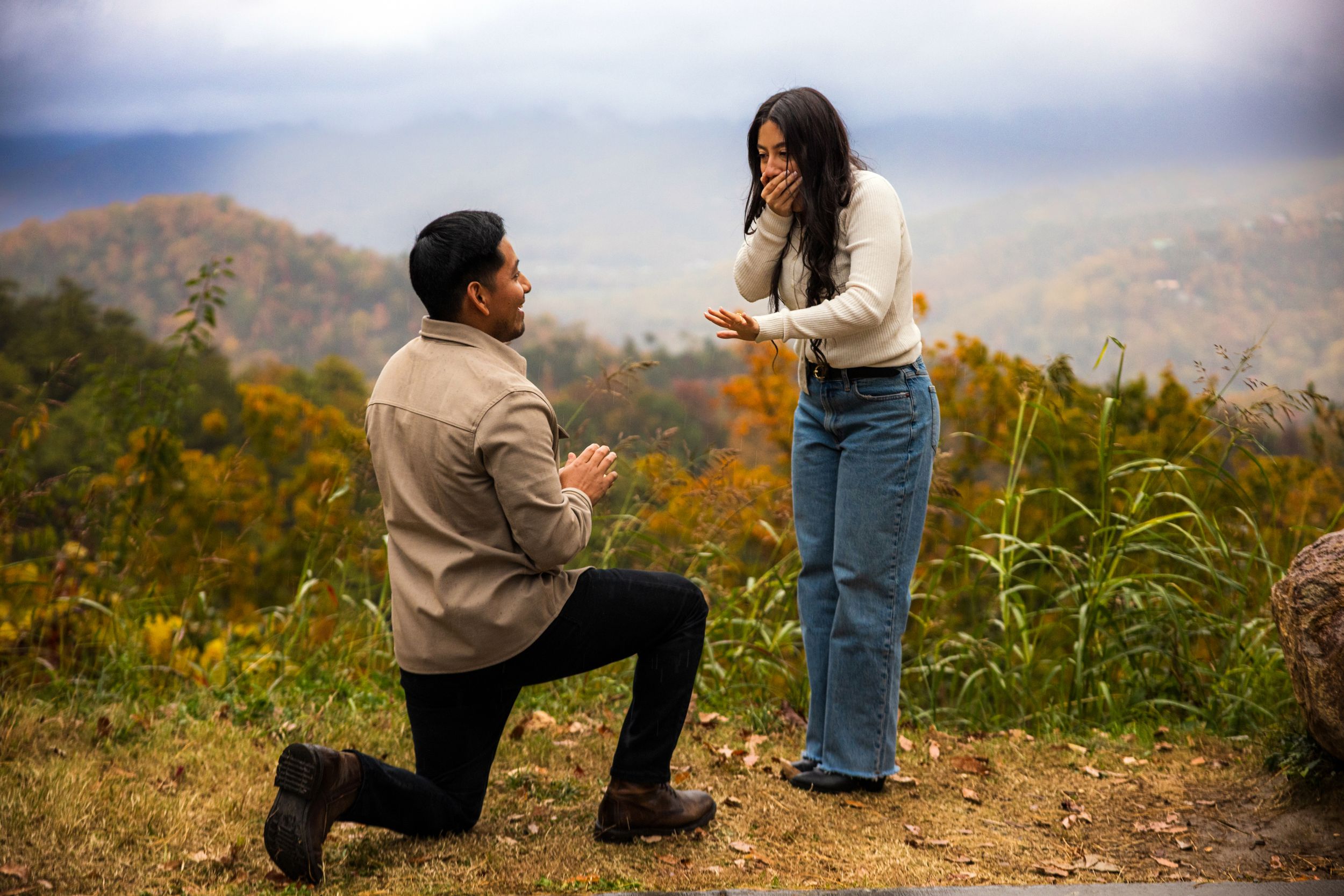 A romantic marriage proposal takes place on a scenic mountaintop during autumn with colorful fall foliage in the background.