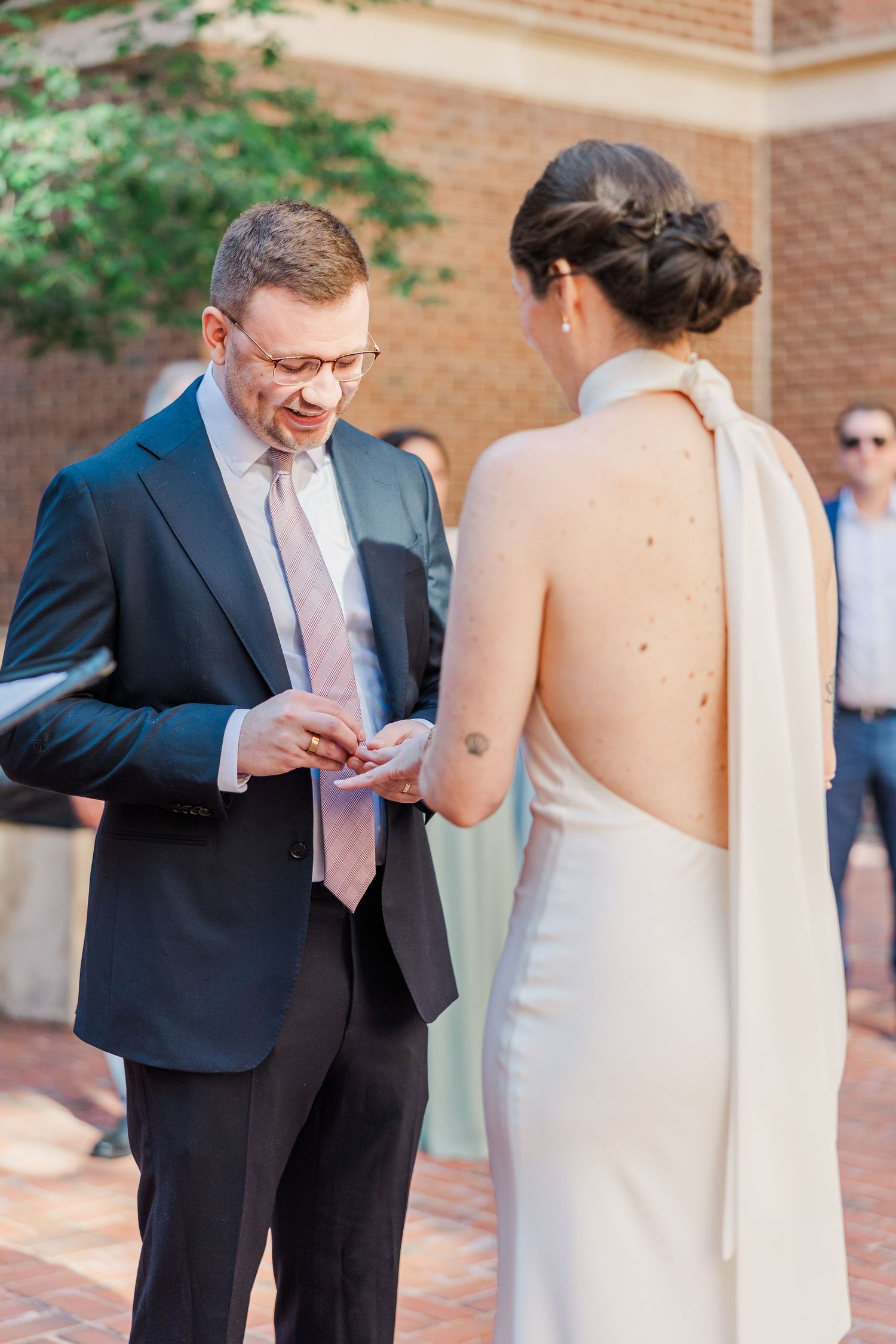 Groom in navy suit placing wedding ring on bride in elegant backless white halter gown during outdoor ceremony.