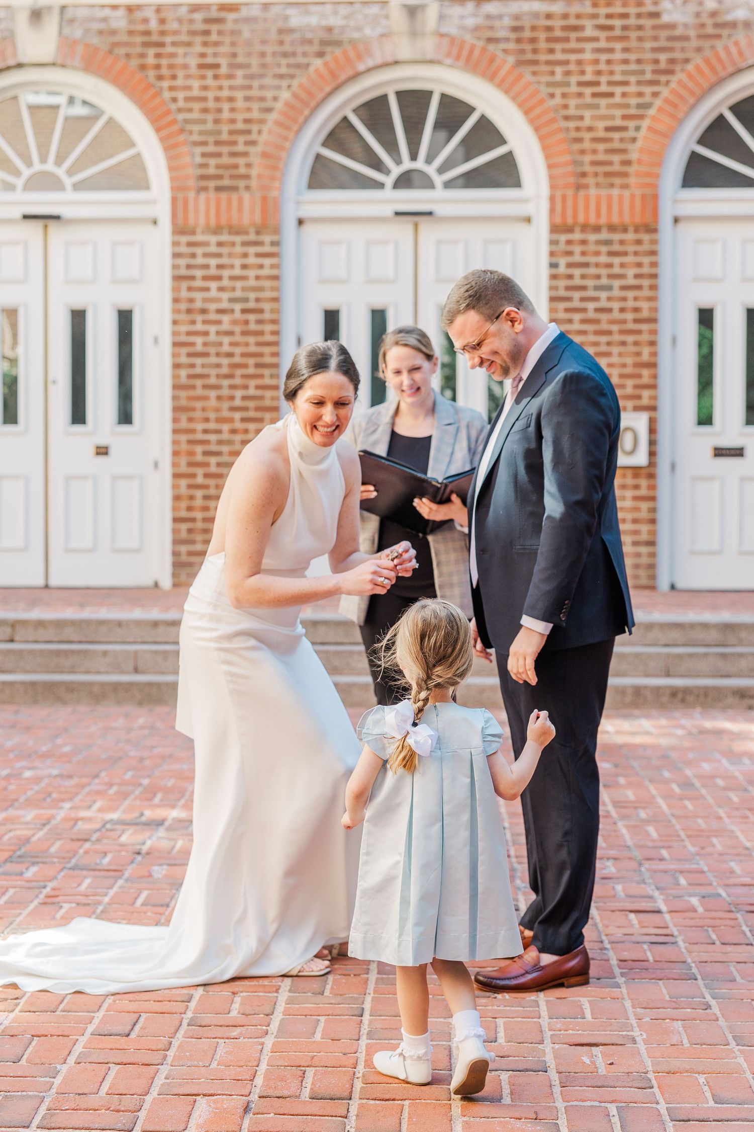 Bride and groom exchange vows outdoors on brick plaza with flower girl and officiant before historic brick building.