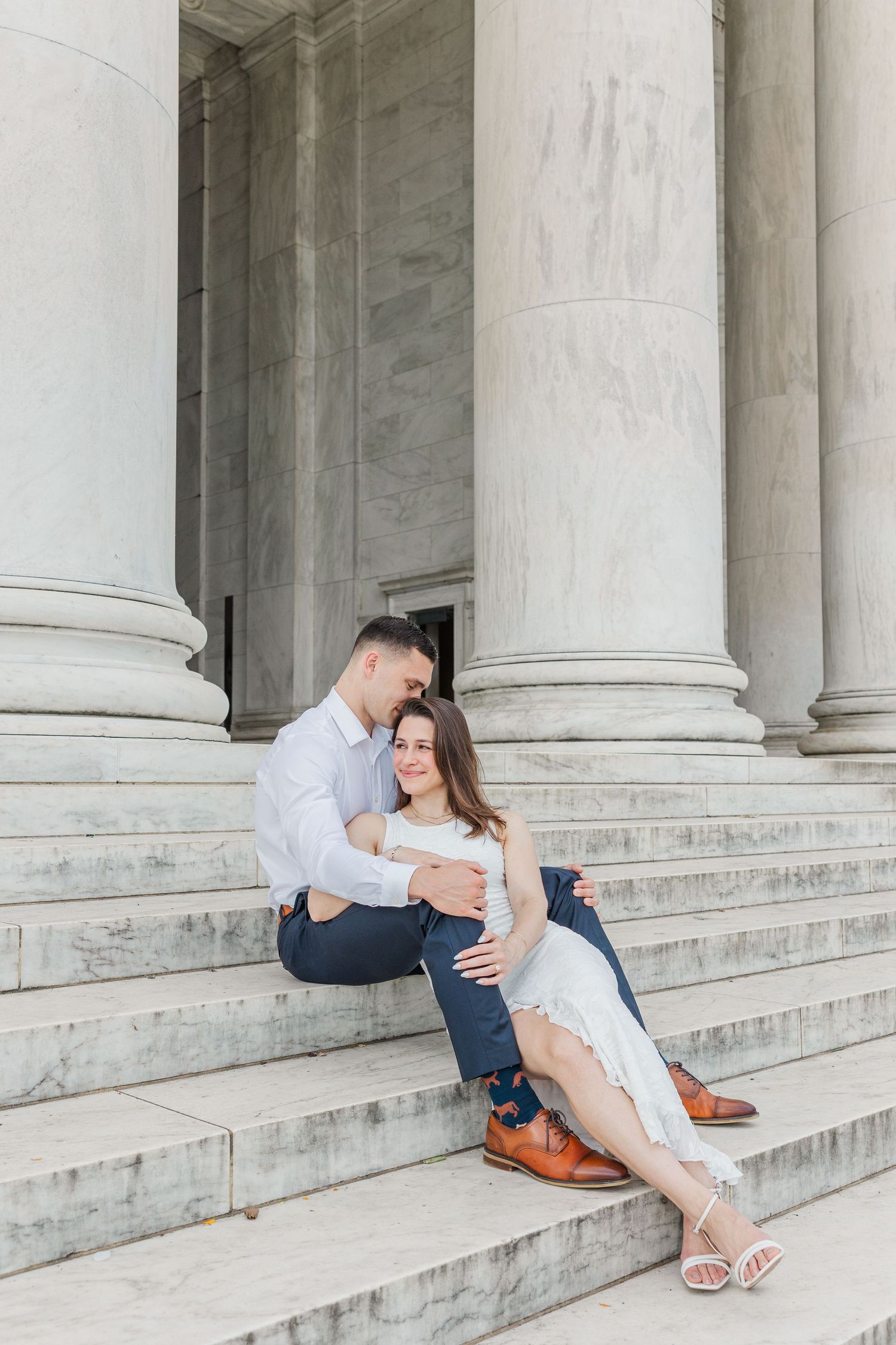 Couple sitting on marble steps of a neoclassical building with columns, embracing tenderly during an engagement photo session.