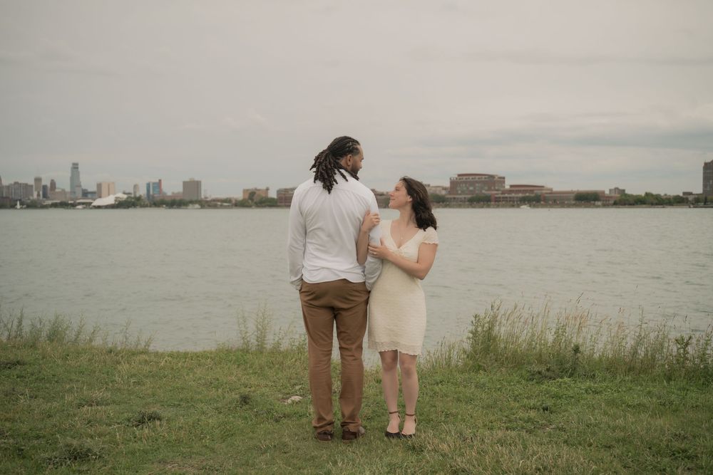 Couple standing together on grass by waterfront with city skyline in background on cloudy day.