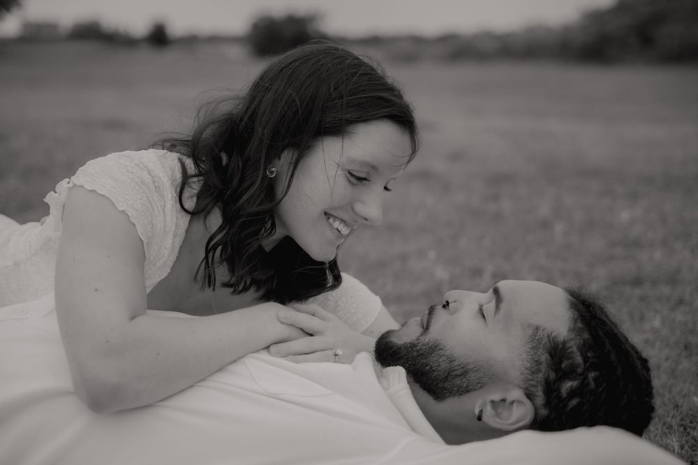 Black and white photo of a couple lying in a field, smiling at each other in a romantic outdoor setting.
