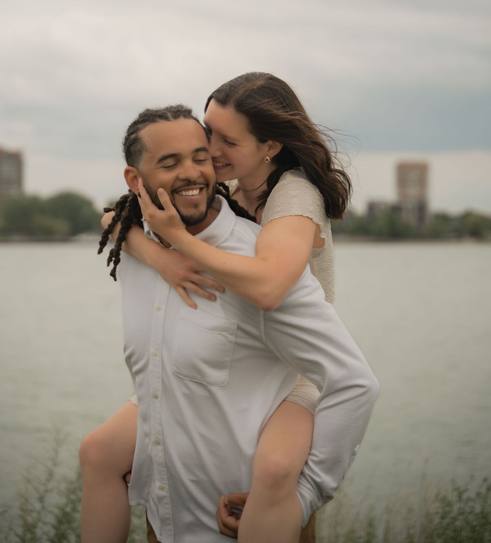 Couple sharing a joyful piggyback moment by a lake, woman smiling and embracing man with dreadlocks in white shirt.