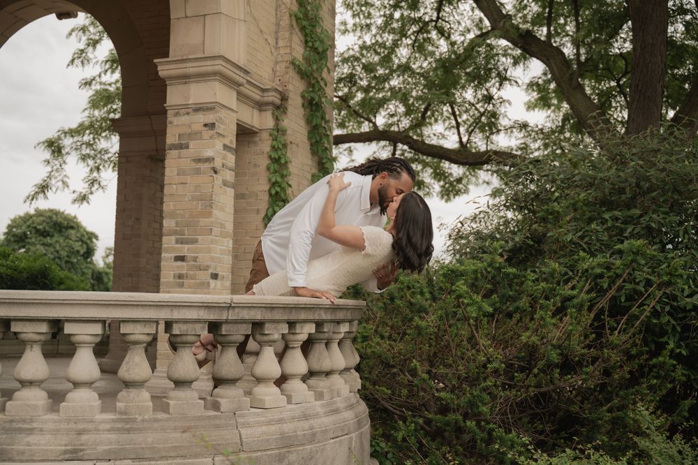 Bride and groom sharing a romantic dip kiss on a stone balcony surrounded by lush greenery at an elegant outdoor venue.