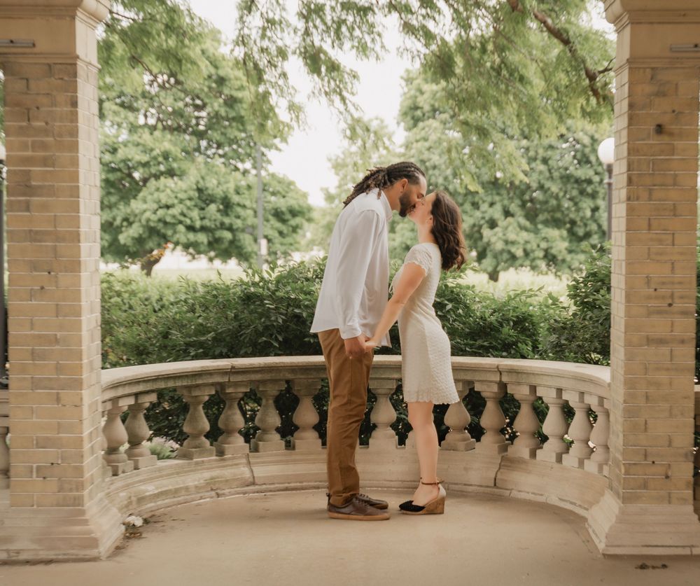 A couple shares a romantic kiss on a stone balcony surrounded by lush greenery and elegant columns.